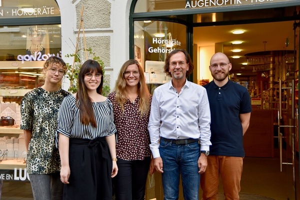 Fünf Mitarbeiter eines Hörgeräte- und Optikfachgeschäfts stehen lächelnd vor dem Laden.